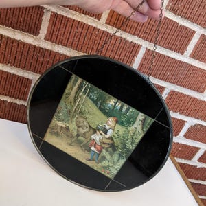 May include: A round, black framed decorative piece with a chain for hanging. The center features a colorful illustration of Little Red Riding Hood and the wolf in a forest scene. The frame has a geometric design.