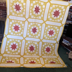May include: A handmade quilt with a repeating pattern of red starbursts on a white background. The stars are surrounded by yellow and white geometric shapes. The quilt is hanging from a clothesline.