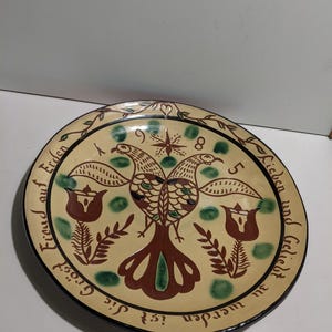 May include: A round ceramic plate with a cream-colored glaze featuring a hand-painted design of two birds, flowers, and green spots. The text reads: 1985 Lieben und Geliebt zu werden ist die Gröst Freud auf Erden, with a vine border.