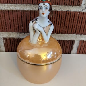 May include: A vintage porcelain trinket box featuring a seated woman figurine. The woman has a painted face, a colorful headpiece, and floral accents on her shoulders. The box is a peach color with a gold trim.
