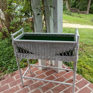 May include: A rectangular white wicker plant stand featuring a woven lattice pattern on the sides and a solid green metal liner. The stand is supported by four vertical legs connected by a horizontal support bar near the base for stability.