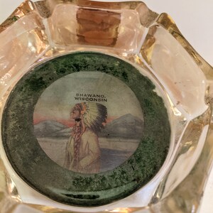 May include: A clear glass ashtray with a round, green-rimmed inset featuring a vintage illustration of a Native American person wearing a headdress. The text "SHAWANO, WISCONSIN" is printed on the inset.