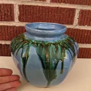 May include: A light blue ceramic vase with a round body and a wide opening. The vase features a green and gold glaze that drips down the sides, creating a unique decorative effect. The vase is a decorative home decor item.