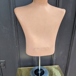 May include: A vintage, tan-colored mannequin torso with a round neck and shoulders. The mannequin is mounted on a chrome pole and a circular base. The base is set on a gold-colored, textured surface. The mannequin is designed for displaying clothing.
