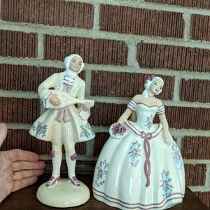 May include: Two ceramic figurines depicting a man playing a lute and a woman in a ball gown. The figurines are cream-colored with pink and blue floral accents and trim. The man is wearing a cream-colored coat and the woman is holding a bouquet.