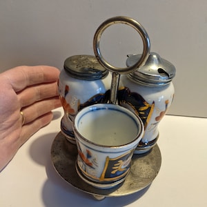 Könnte beinhalten: Ein vintage Salz- und Pfefferstreuer-Set aus Porzellan mit passendem Gewürzhalter. Das Set hat ein blaues und weißes Design mit goldenen Akzenten. Die Streuer haben silberne Deckel mit einem kleinen Loch zum Ausgießen.