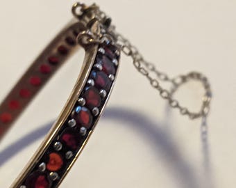 Puede incluir: Primer plano de una pulsera de brazalete dorada con una fila de pequeñas piedras preciosas rojas y redondas. La pulsera tiene un cierre de bisagra y una delicada cadena. Las piedras están engastadas en un metal oscuro.