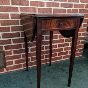 May include: A dark brown, antique drop-leaf table with a single drawer and a round pull. The table has four slender legs and a curved apron. The table is made of wood and has a polished finish.