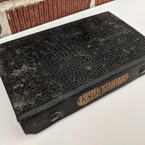 Pu&ograve; includere: Una custodia rettangolare nera vintage con un motivo strutturato simile a quello di un rettile. La custodia ha un'etichetta dorata con la scritta "KNU EYEGLASS MOUNTING". Sono visibili due piccoli rivetti rotondi color argento.