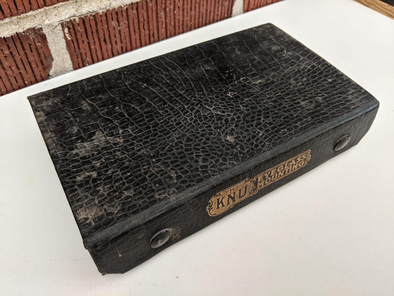 Pu&ograve; includere: Una custodia rettangolare nera vintage con un motivo strutturato simile a quello di un rettile. La custodia ha un'etichetta dorata con la scritta "KNU EYEGLASS MOUNTING". Sono visibili due piccoli rivetti rotondi color argento.