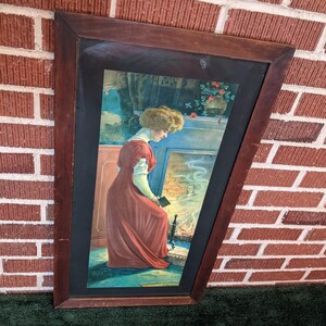May include: A framed vintage print of a woman in a red dress standing in front of a fireplace. The woman is holding a book and looking down at it. The fireplace is lit and there is smoke coming from it. The print is in a dark wood frame.