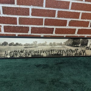 May include: A long, rectangular black-framed panoramic photograph in black and white. The image depicts a large group of people standing in rows outdoors on a grassy field, with trees and a vintage vehicle visible in the background.
