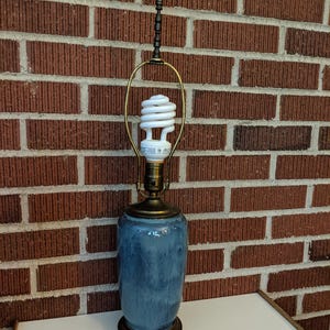 May include: A table lamp featuring a cylindrical ceramic base with a blue and grey mottled glaze. The lamp has a brass-toned metal base and hardware, a metal harp, and a white spiral compact fluorescent light bulb topped with a blue finial.