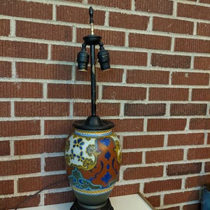 May include: A table lamp featuring a ceramic base with a colorful pattern of blue, orange, yellow, and green floral motifs. The lamp has a black metal stem with two light sockets, a black finial, and a dark circular base. A brown power cord is attached.
