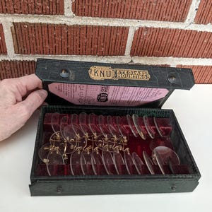 May include: An open, vintage black case with a red velvet interior, holding multiple clear eyeglass lenses with gold-colored frames. The case is labeled "KNU Eyeglass Mounting" in gold lettering. The background is a red brick wall.