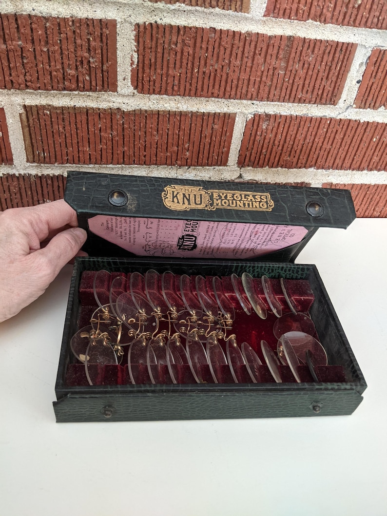 Pu&ograve; includere: Una custodia nera vintage aperta, contenente pi&ugrave; lenti per occhiali trasparenti con montature color oro. La custodia &egrave; etichettata "KNU Eyeglass Mounting" in lettere dorate. Lo sfondo &egrave; un muro di mattoni rossi.
