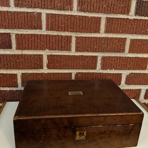 May include: A rectangular wooden box with a dark brown burl wood grain finish. The box features a small, rectangular metal plaque centered on the lid and a brass-colored keyhole escutcheon plate centered on the front panel of the closed lid.