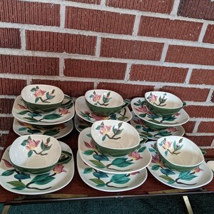 Könnte beinhalten: Ein Set aus 10 Vintage-Teetassen und Untertassen mit einem Blumendesign. Die Tassen und Untertassen sind weiß mit grünen Blättern und rosa Blüten. Das Set ist auf einem Holztisch gestapelt.