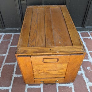 May include: A vintage wooden storage cabinet with a warm, natural wood finish. The cabinet features a hinged top and a drawer with a metal pull. The wood grain is visible, adding to its rustic charm. The cabinet is sitting on a brick surface.