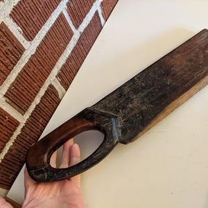 May include: A vintage wooden drawknife with a dark, weathered appearance. The blade is rectangular, and the handle is a dark brown with an oval opening for grip. The tool is likely used for shaping wood.