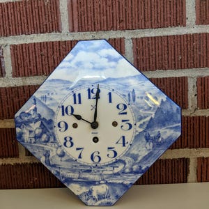 May include: A blue and white ceramic wall clock with a landscape design. The clock face has black hands and blue numerals. The clock is octagonal and mounted on a red brick wall.