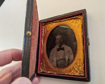 May include: Antique portrait photograph in a decorative case. The oval portrait features a man in a suit and bow tie. The case is black with a gold-colored frame around the portrait and a red velvet interior.