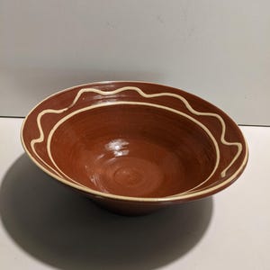 May include: A round, terracotta-colored ceramic bowl with a glossy finish. The interior and exterior feature a single, continuous cream-colored wavy line pattern that encircles the upper rim of the bowl, creating a distinct contrast.