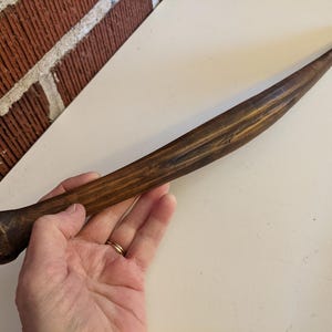May include: A handcrafted wooden rolling pin with a dark brown finish. The rolling pin has a rounded handle and tapers to a point. The wood grain is visible, adding to its rustic charm. A kitchen utensil for baking.