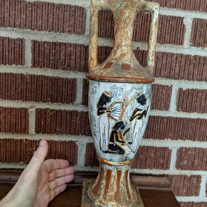 May include: A ceramic vase with a white background and gold accents. The vase features a design of three figures playing musical instruments. The vase is approximately 12 inches tall.