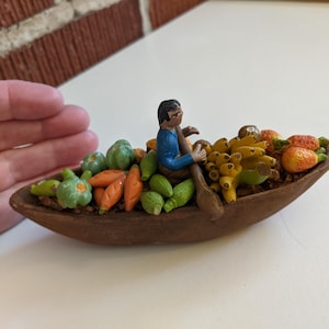 May include: A small brown boat figurine carrying a person in a blue shirt and brown pants who is holding a wooden paddle. The boat is filled with a variety of colorful miniature produce including green vegetables, orange carrots, and yellow bananas.