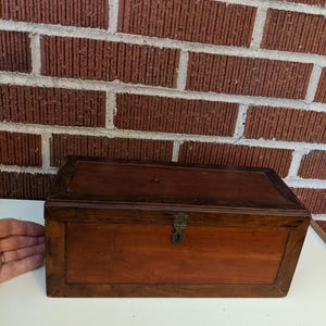 May include: A rectangular wooden box with a dark brown frame and a lighter brown center panel. The box has a metal clasp and a small keyhole. The background is a red brick wall.
