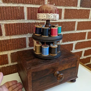 May include: A vintage sewing kit with a dark wooden box base and a rotating thread spool holder. The spool holder has multiple spools of colorful thread and a pin cushion on top. The box has a drawer with a wooden knob.
