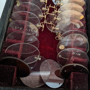 Pu&ograve; includere: Una collezione di occhiali e lenti antichi, esposta in una custodia nera con interno in velluto rosso. Le lenti trasparenti, di forma ovale, presentano scritte come "STIC-TITE". Le montature sono dorate.