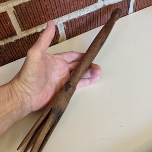 May include: A vintage wooden fork with a long handle and four tines. The fork is a dark brown color and appears to be handmade. The handle has a decorative carving near the top. The fork is held in a hand.