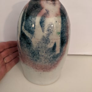 Puede incluir: Un jarrón de cerámica blanca con cuerpo redondeado y cuello estrecho. El jarrón está decorado con patrones abstractos en tonos azules, rosas y verdes.
