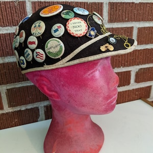 May include: A black felt baseball cap with a white brim and a white stripe around the crown. The cap is covered in vintage buttons and pins, including a button that says "I Never Noo That".