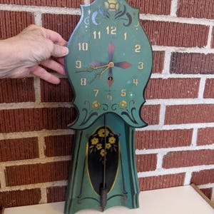May include: A vintage green and black wooden clock with gold accents. The clock features a floral design and a pendulum. The clock face has gold numerals and hands. The clock is approximately 24 inches tall.