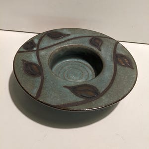 May include: A round ceramic candle holder with a teal and blue mottled glaze. The wide, flat rim features a dark brown hand-painted vine pattern with leaves encircling the center. The interior base has concentric circular grooves.