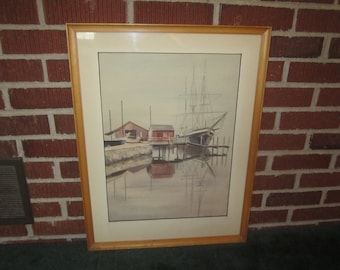 May include: A framed watercolor painting depicting a ship docked at a pier with two small red buildings in the background. The water is calm and reflects the sky and the ship. The painting is in a light wood frame.