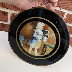 May include: A round, black decorative plate with a gold border and a painted scene of a person in a blue coat and white wig. The plate is held by a chain and has a brass-colored rim.