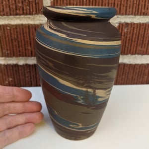 May include: A decorative ceramic vase with a dark brown base and a blue rim. The vase features a swirled pattern of brown, blue, and beige stripes. The vase is cylindrical with a slightly flared top.