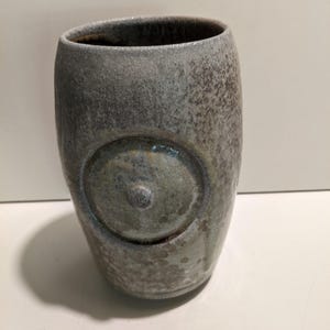 May include: A cylindrical ceramic tumbler with a mottled gray and brown glaze. The exterior features a prominent circular relief element centered on the front, with a small raised dot in the middle of the circle, creating a textured appearance.