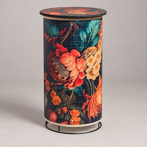 May include: A cylindrical side table featuring a dark background adorned with a vibrant floral pattern of large orange, red, and cream-coloured flowers and green leaves. The table has a flat circular top and a thin black metal base frame.