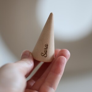 May include: A light-colored, cone-shaped wooden ring holder. The word "Sara" is engraved on the side of the holder. The holder is held in a person's hand.