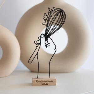 Könnte beinhalten: Schwarze Drahtskulptur einer Hand, die einen Schneebesen hält, mit dem Namen "Nathalie" darüber. Die Skulptur ist auf einem Holzsockel mit dem Text "Konditormeisterin 2026" montiert. Beige Vasen im Hintergrund.