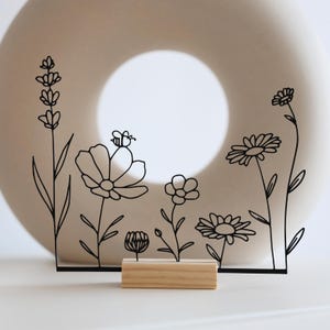 Könnte beinhalten: Schwarzes Metall-Blumendesign mit Holzsockel. Das Design zeigt verschiedene Blumen und eine Biene. Der Hintergrund ist ein cremefarbenes, kreisförmiges Objekt. Das Design ist wahrscheinlich ein dekorativer Artikel.