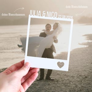 Peut inclure: Un cadre blanc rectangulaire découpé rappelant une photo instantanée vintage, avec le texte JULIA & NICO 22.08.2026 en haut et une découpe en forme de cœur dans le coin inférieur droit, conçu pour présenter une photographie de mariage.