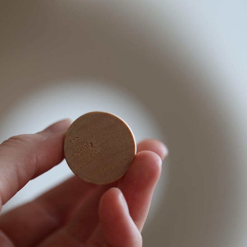 May include: A small, round, light brown object is held between fingers. The object has a slightly textured surface and appears to be made of a natural material. The background is a soft, blurred neutral tone.
