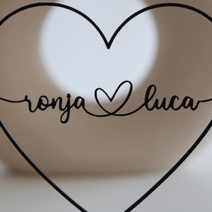 May include: Black metal heart-shaped wall decor with the names "ronja" and "luca" in cursive script. A heart symbol connects the names. The design is minimalist and suitable for home decoration.