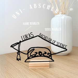 May include: A black graduation cap decoration with the name "Lukas", the title "Bachelor of Science", and the year "2025". The decoration is mounted on a small wooden block. A white vase with dried flowers is in the background.